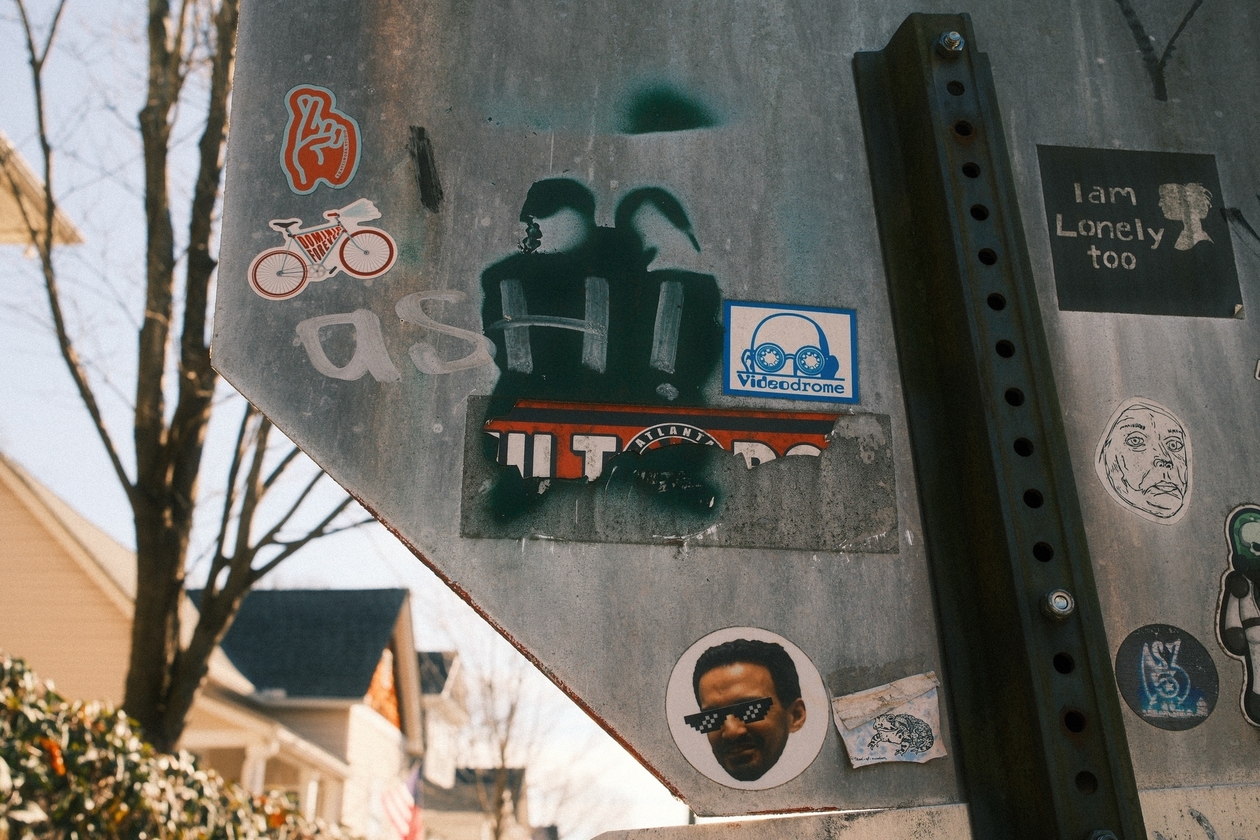 A street sign covered with various stickers and graffiti. The stickers include a hand with crossed fingers, a red bicycle, a labeled “Videodrome” with goggles, a face wearing shaded sunglasses, and a text stating “I am Lonely too.