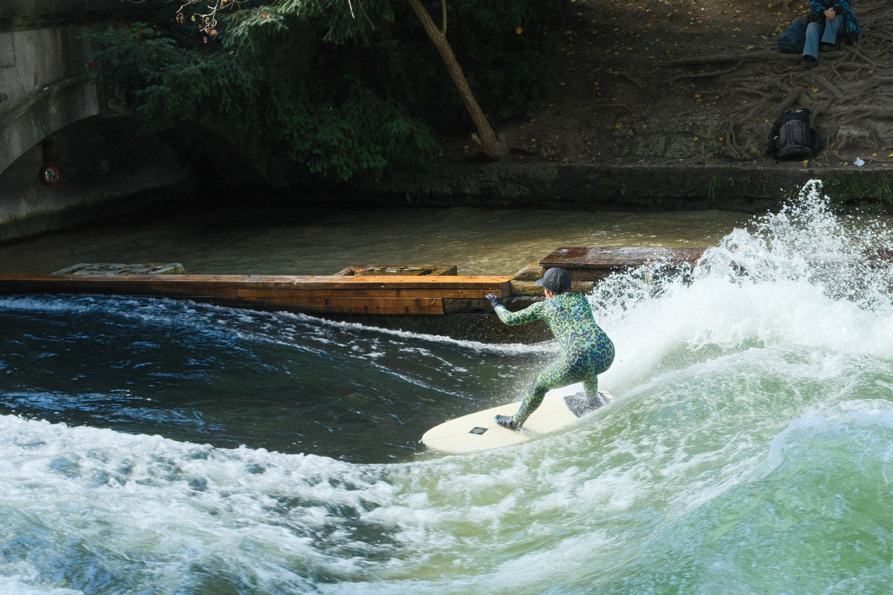 A person in a wetsuit is surfing on a wave in a river, with onlookers in the background.