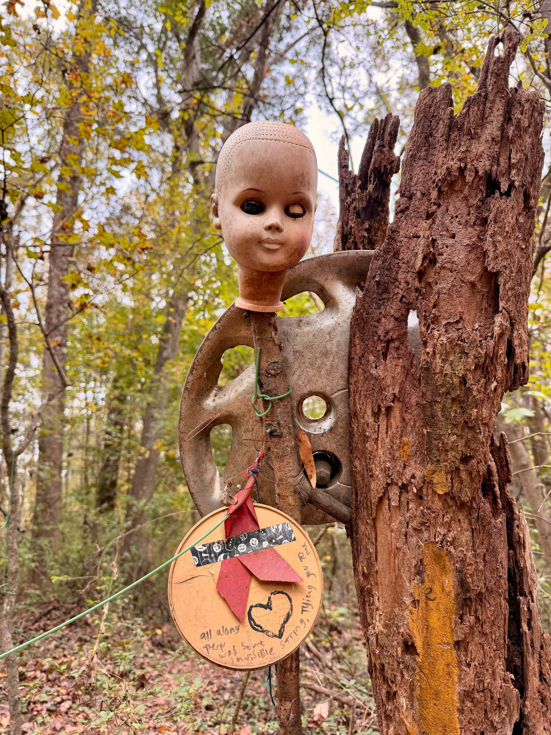 Auto-generated description: A doll&rsquo;s head is mounted on a rusted wheel attached to a tree stump, with a wooden disc featuring drawings and notes hanging below, set against a forested background.