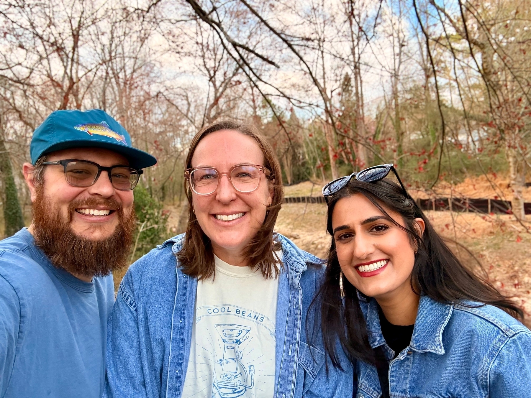 Three people are smiling in an outdoor setting with leafless trees in the background. The person on the left has a beard and wears glasses and a blue cap. The person in the middle also wears glasses and a denim shirt. The person on the right has glasses on their head and is also wearing a denim shirt. 