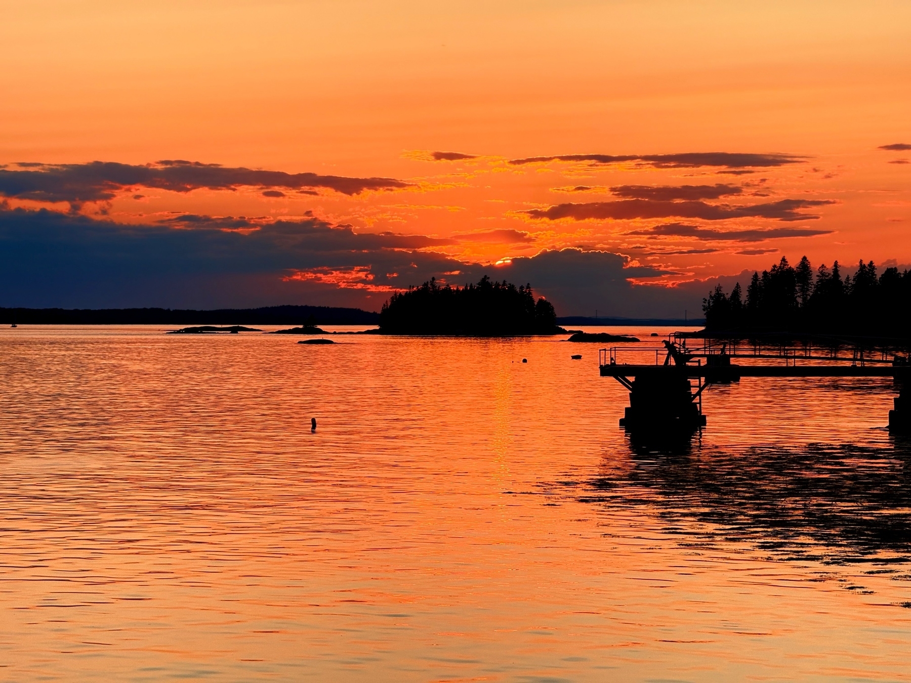 A vibrant sunset over a calm body of water with silhouettes of a dock and trees in the foreground. The sky is illuminated with shades of orange and red, reflecting beautifully on the water&rsquo;s surface.