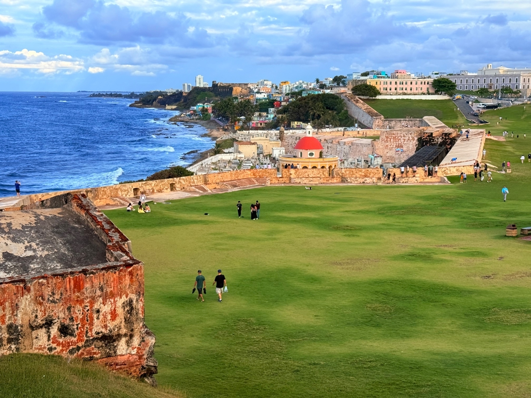 Scenic view of a coastal area with historic fortifications, grassy fields, and a red-domed building in the foreground. People are walking and relaxing on the grass. The ocean is on the left and a vibrant town is visible in the background. 