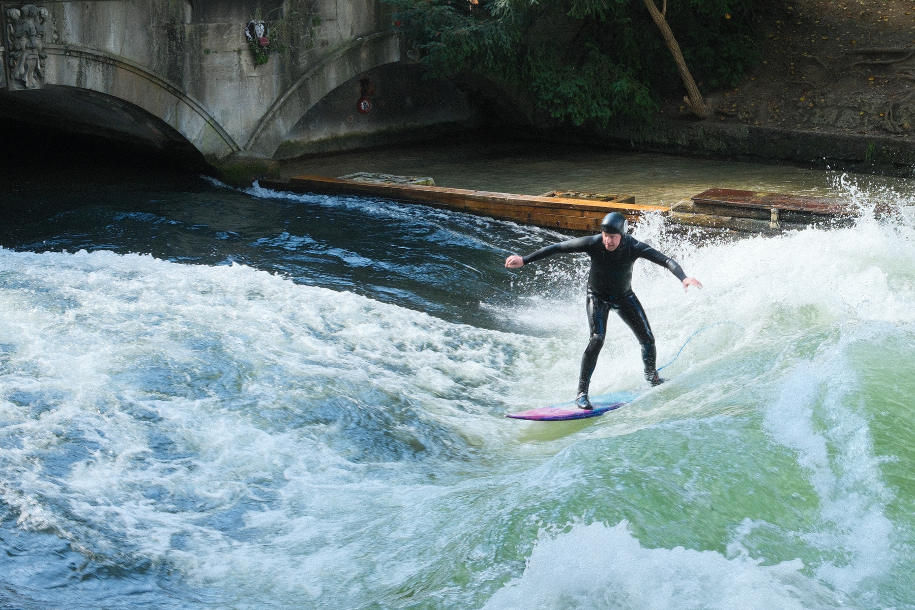 A person is surfing on a standing wave in a river near a bridge.