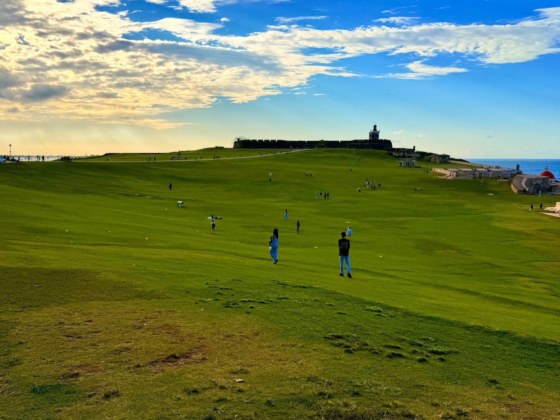 A vast grassy field with several people scattered throughout, under a partly cloudy sky. A historic fort is visible on a hill in the background, with the ocean to the right.
