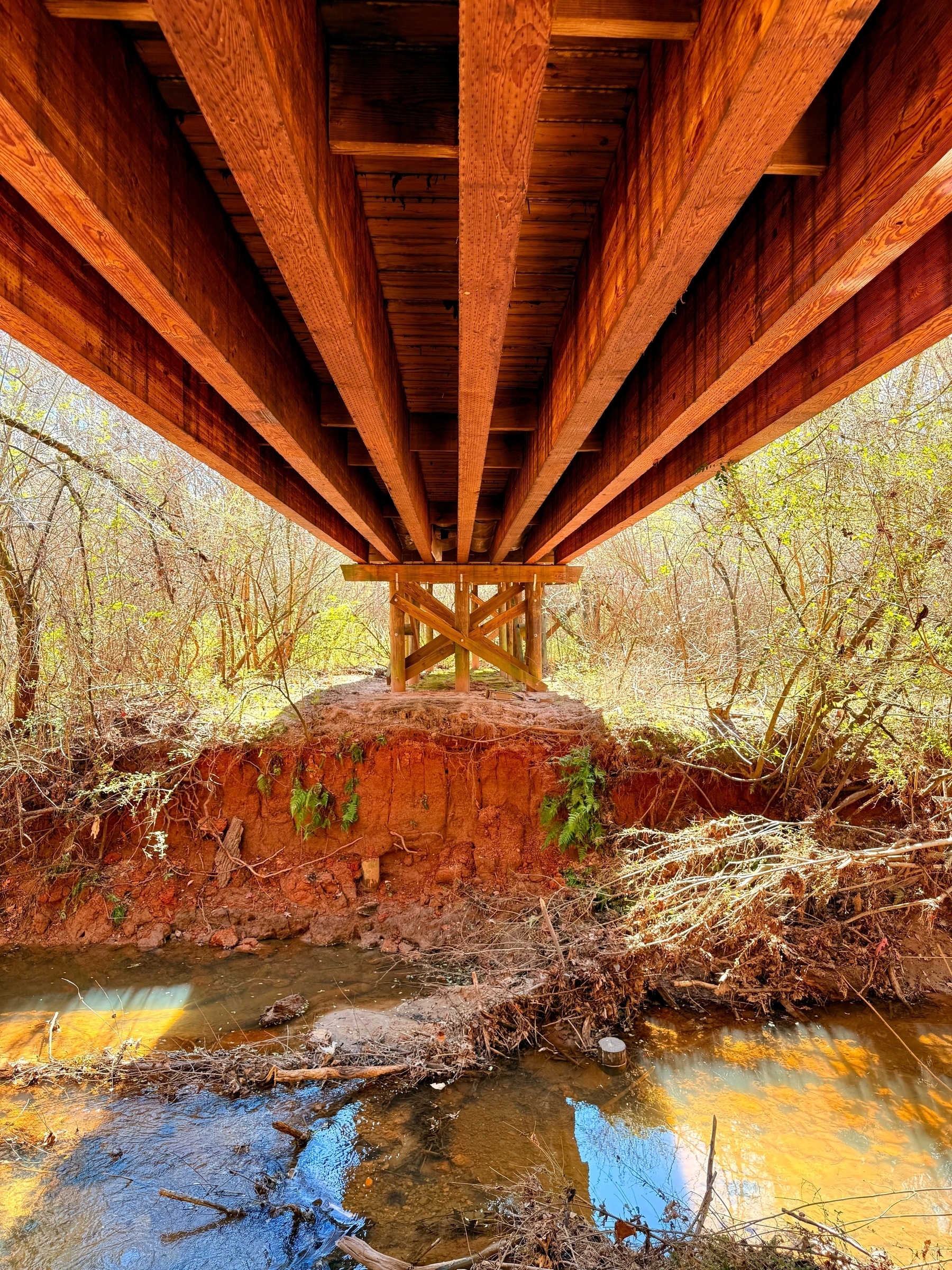 Underneath view of a wooden bridge with beams and supports, spanning over a small creek. The surrounding area features exposed soil and vegetation with sunlight filtering through the branches.