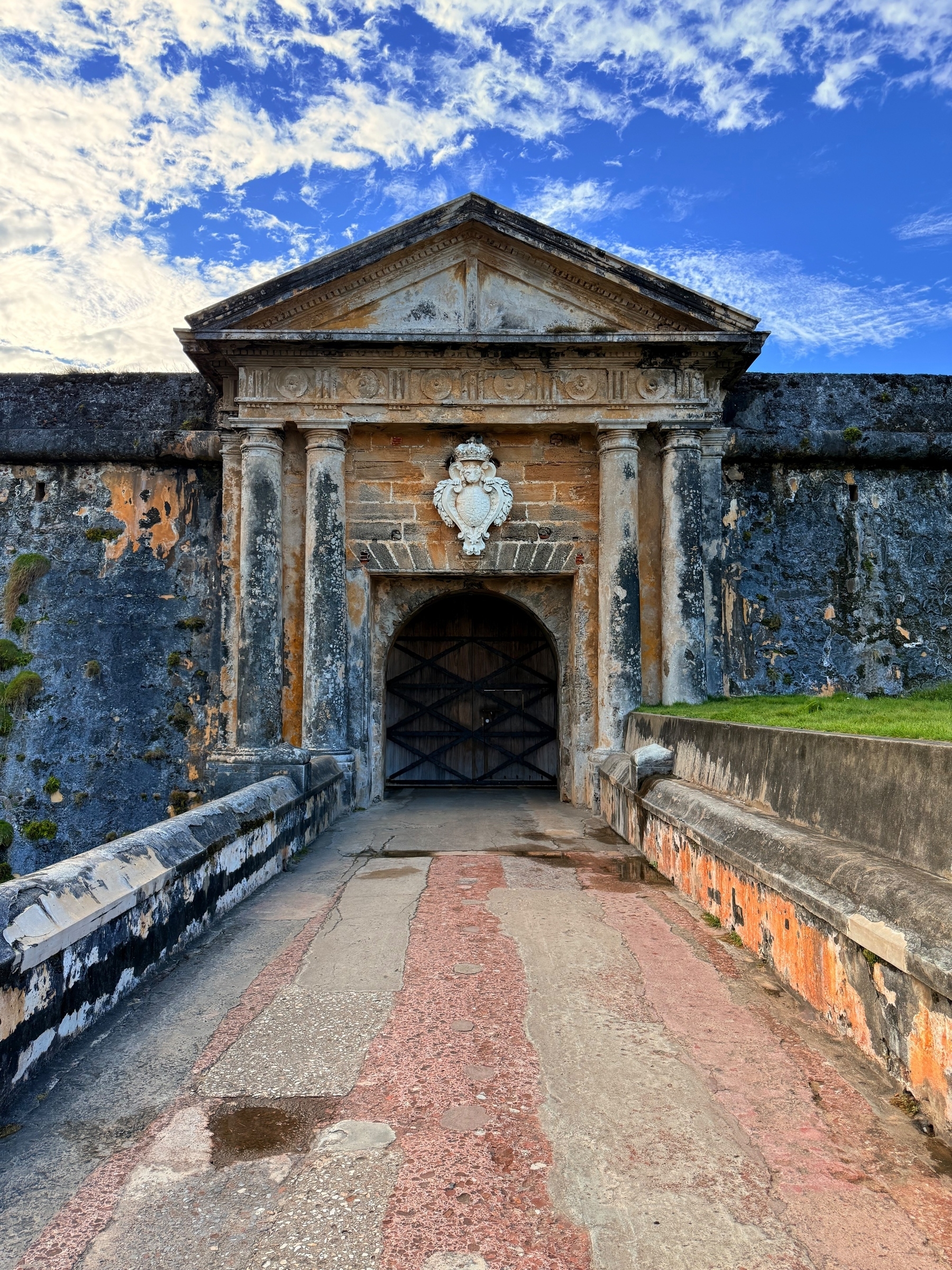 Entrance to an old stone fortress with a wooden gate, featuring classical architectural elements like columns and a carved pediment. The sky is partly cloudy.