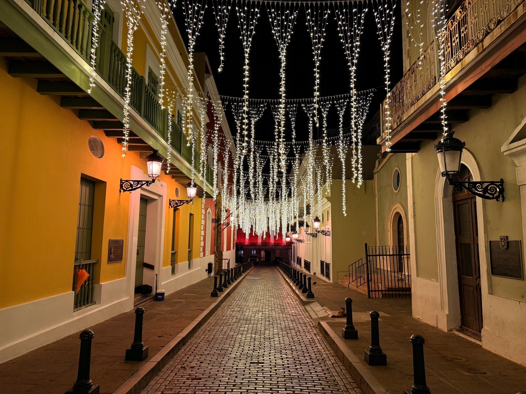 A cobblestone street lined with colorful buildings adorned with ornate balconies. Strings of white lights hang overhead, illuminating the scene at night. Wall-mounted lanterns provide additional warm lighting, creating a festive atmosphere.