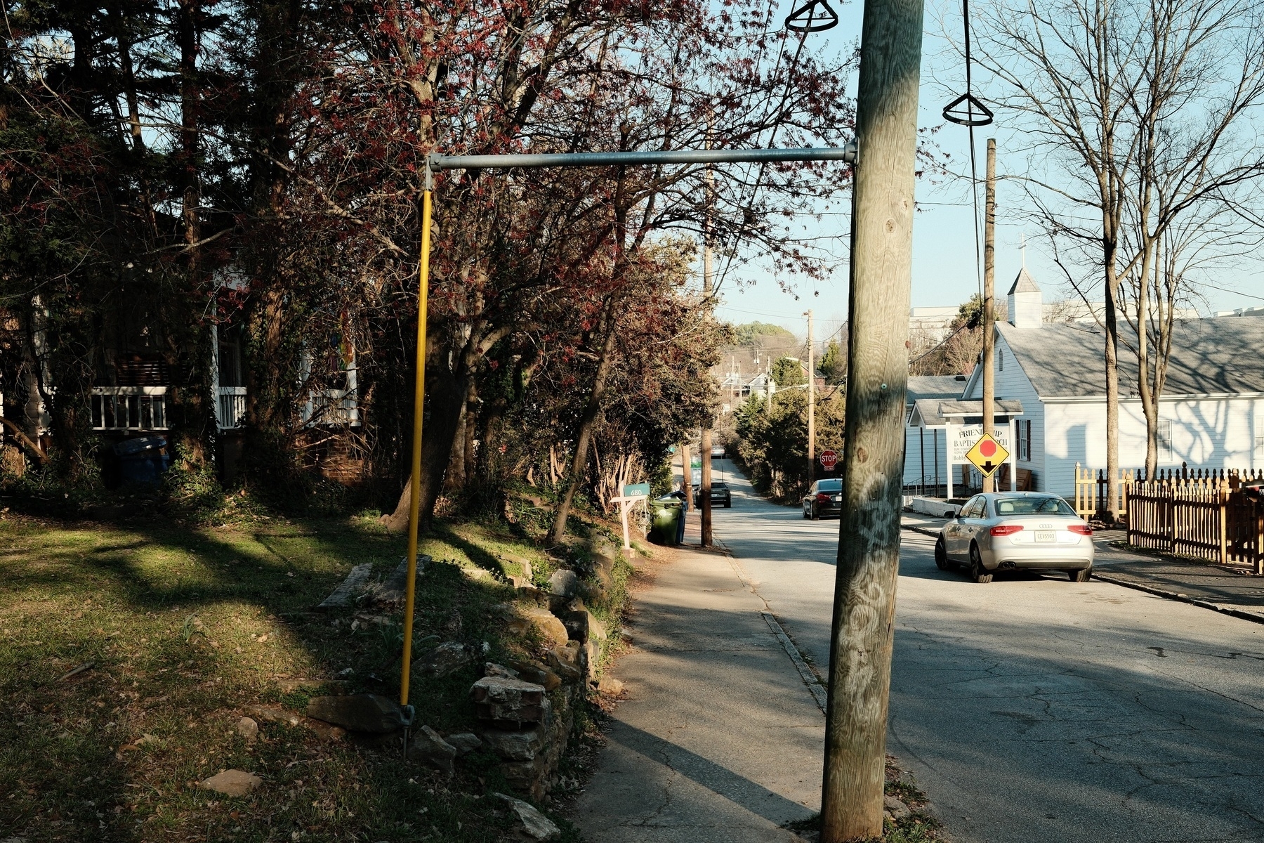 A residential street with parked cars and a stop sign. A wooden utility pole stands by the sidewalk. Trees and bushes line the left side, casting shadows on a grassy area. A white building with a cross on top is visible on the right.