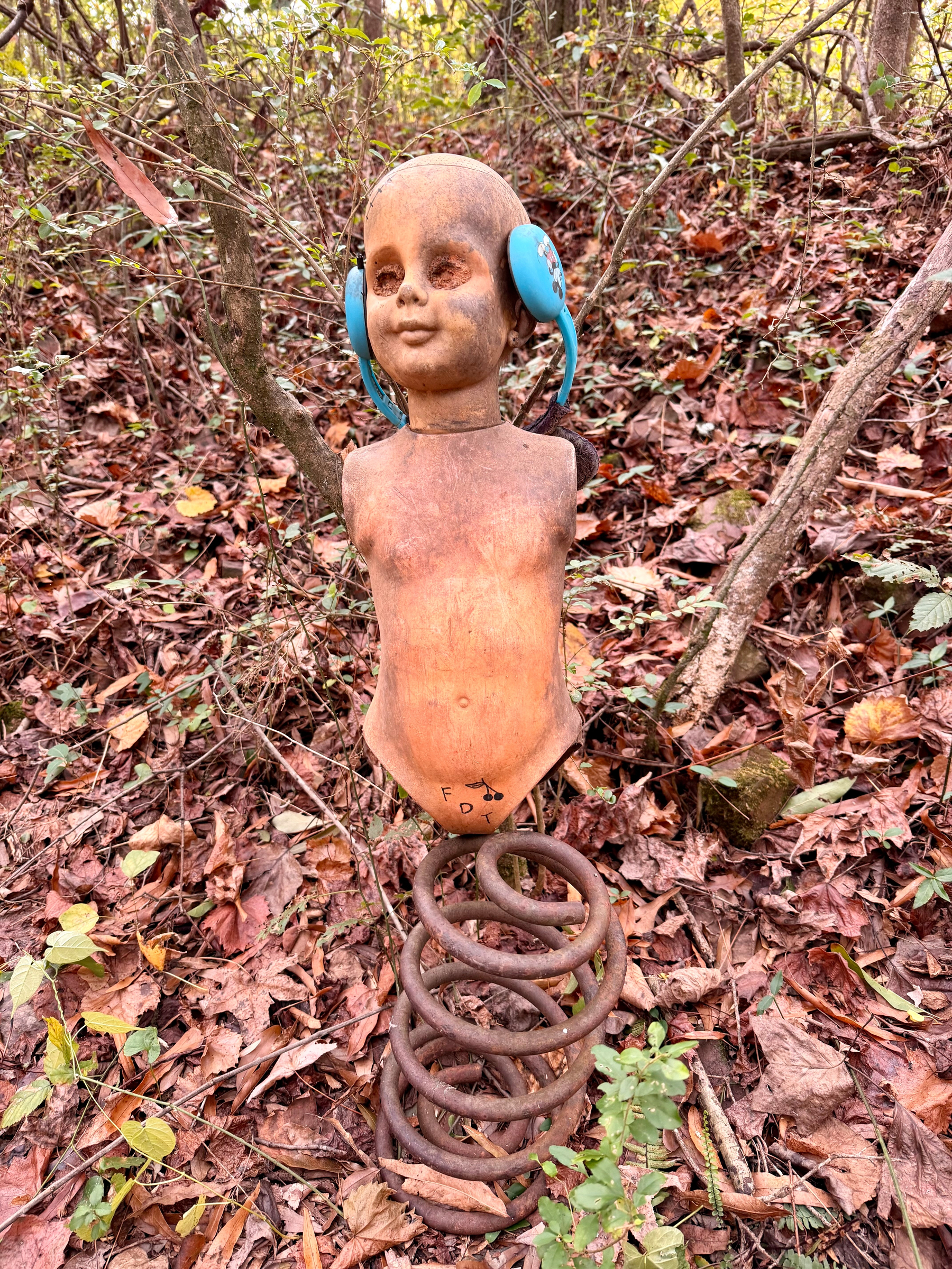 Auto-generated description: A torso of a doll with blue earmuffs is mounted on a metal spring in a forest setting.