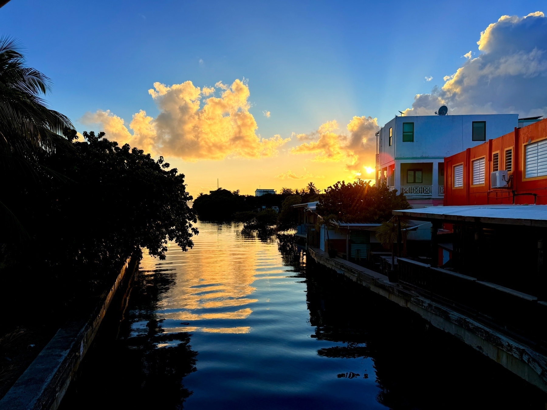 Sunset over a canal with vivid clouds reflecting on the water. Brightly colored buildings line the right side, while lush trees frame the left.