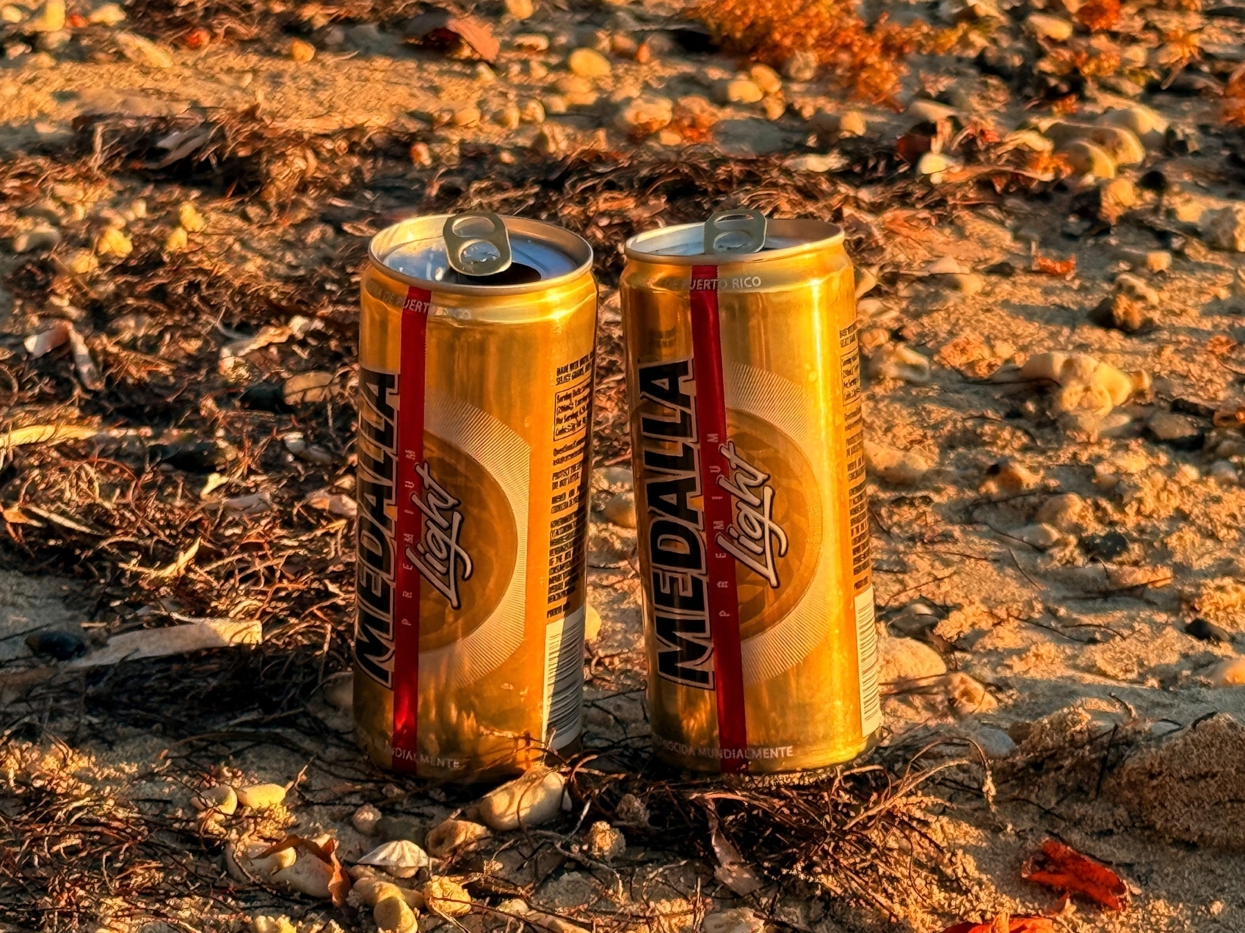 Two cans of Medalla Light beer on sandy ground with small rocks and dried seaweed around them, illuminated by warm sunlight.