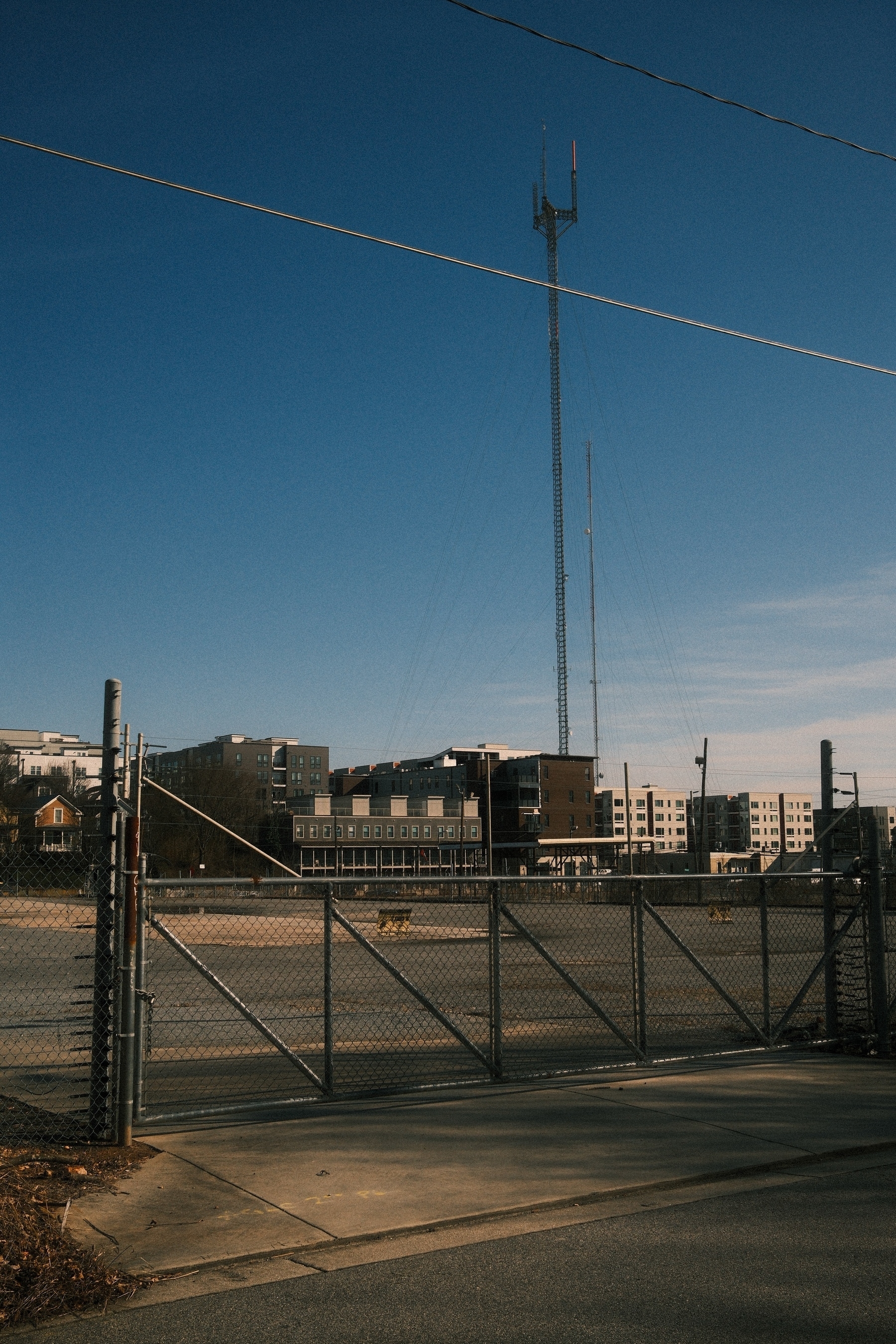 A tall communication tower against a clear blue sky, surrounded by modern apartment buildings. In the foreground, there is a chain-link fence with an open gate, leading to a paved area.
