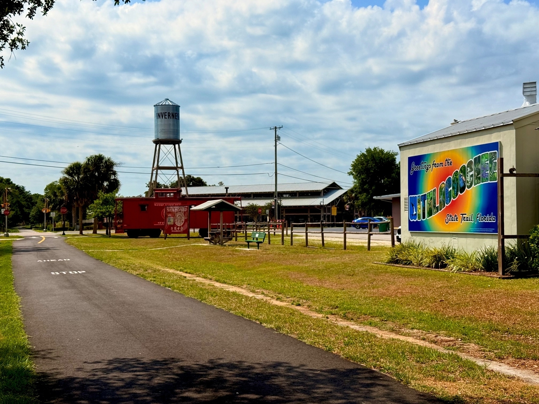 A paved trail with markings runs through a grassy area with a water tower labeled &ldquo;Inverness&rdquo; in the background. There&rsquo;s a red railroad car next to a picnic area with benches and a colorful sign that reads, &ldquo;Greetings from the Withlacoochee State Trail, Florida.”