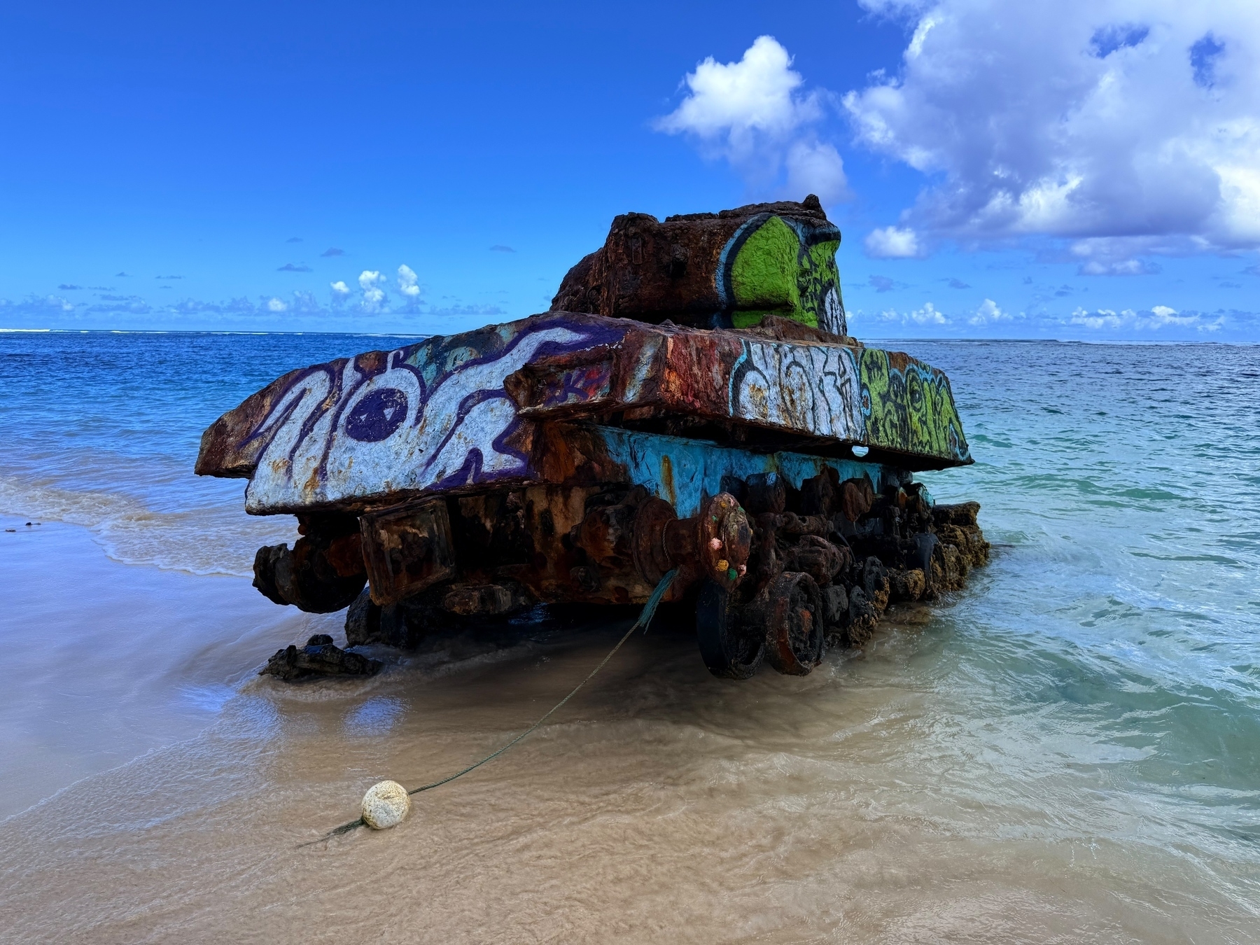 An old, rusted tank sits partially submerged on a sandy beach, covered in colorful graffiti. The blue ocean and a sky with clouds are in the background.