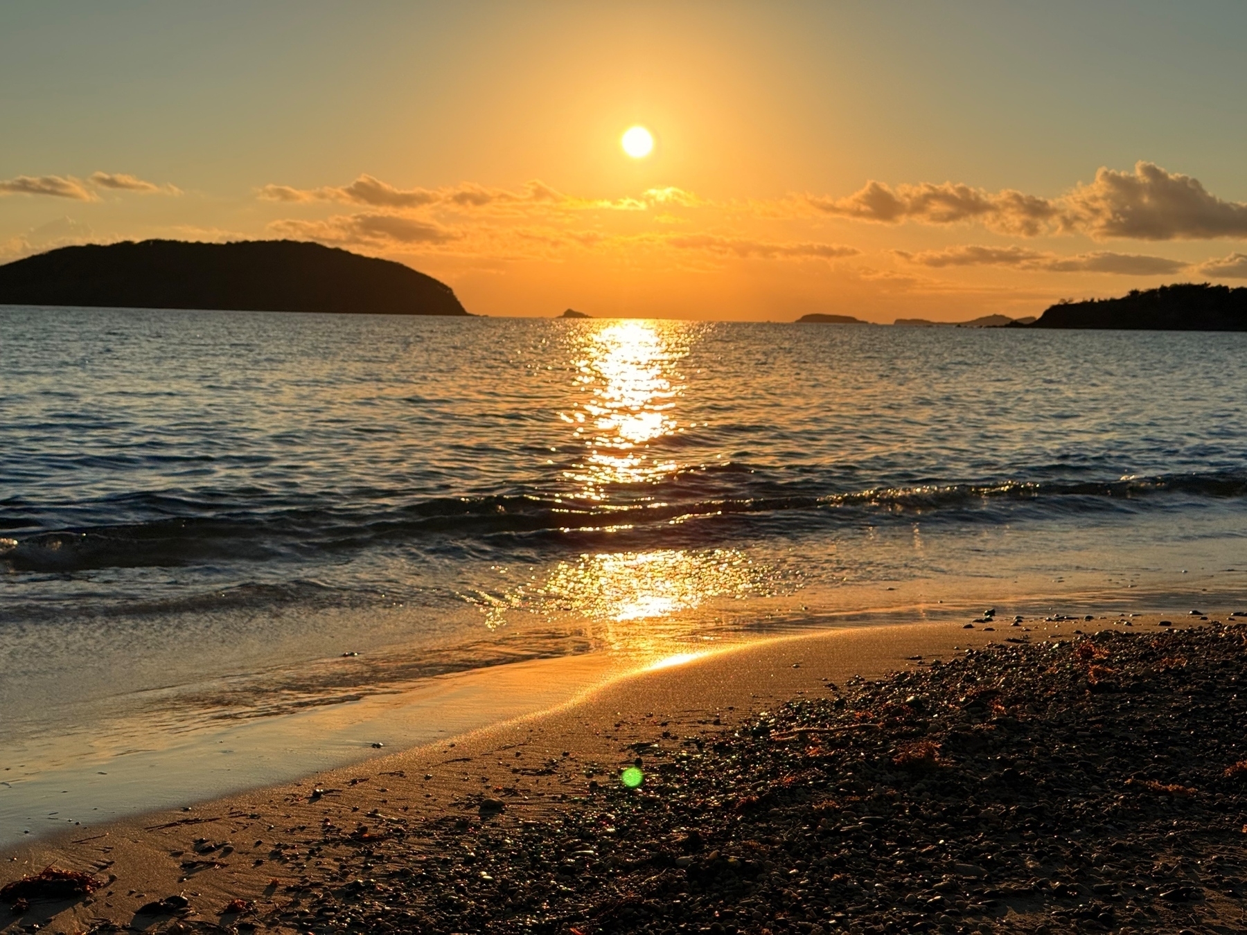 Sunset over the ocean with a glowing sun reflecting off the water. Silhouetted islands in the background and a sandy beach with scattered seaweed in the foreground.