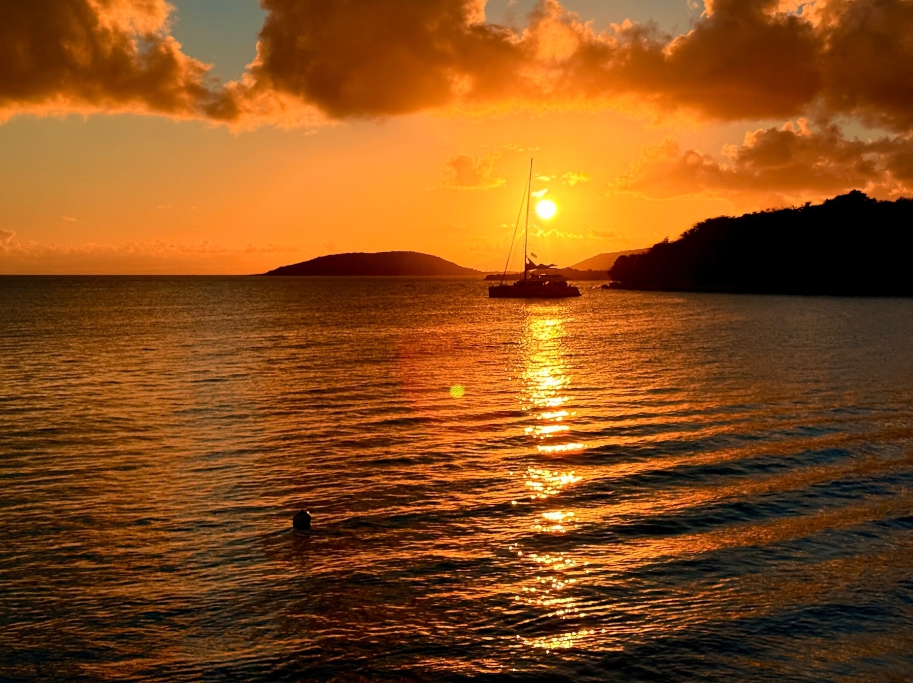 A serene sunset over the ocean with a sailboat silhouetted against the vibrant orange sky. The sun reflects on the water, creating a shimmering path. An island is visible in the background with some clouds adding texture to the sky.