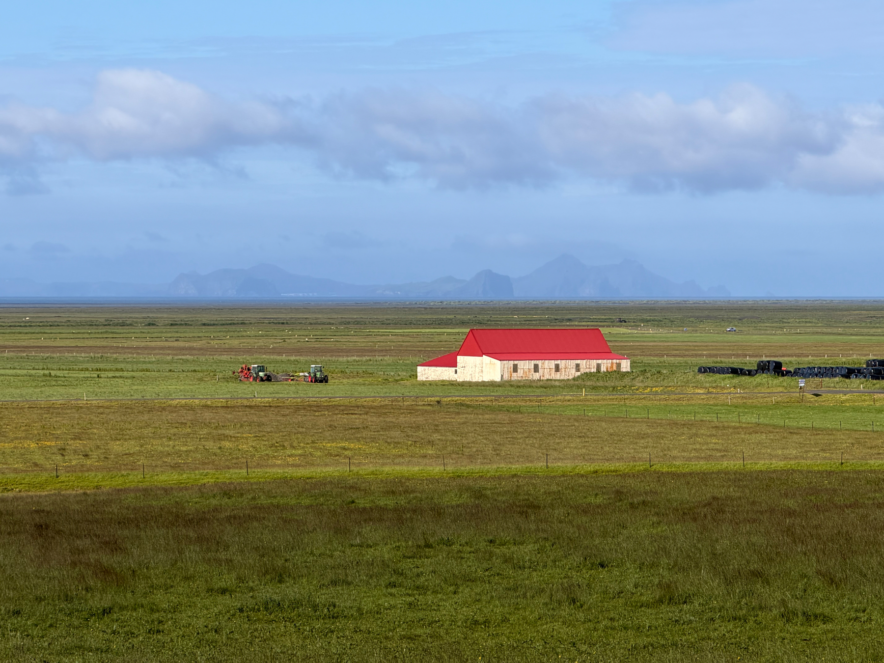 A building with a red roof is situated in the middle of a vast green field with distant mountains visible on the horizon.