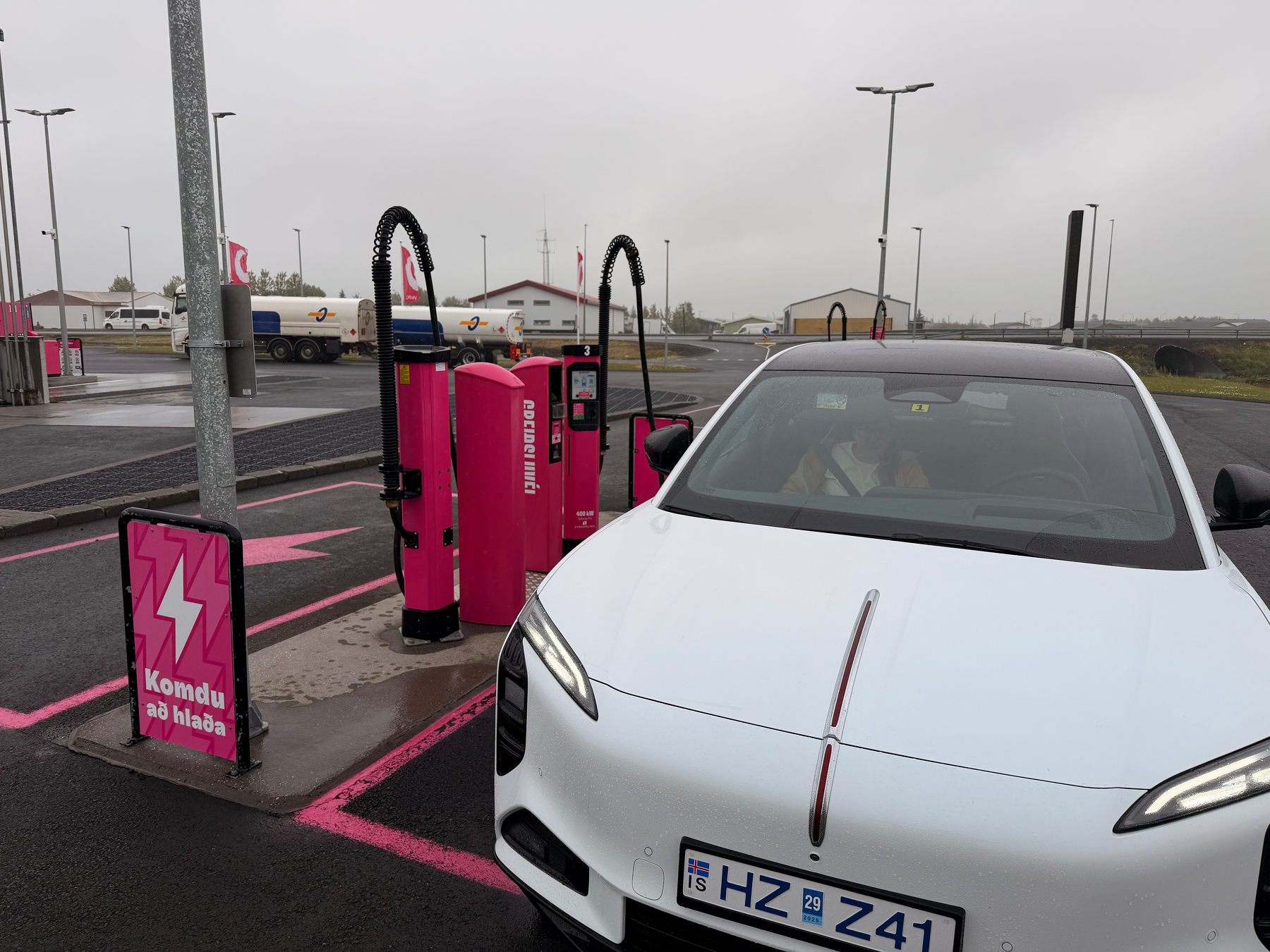 A white electric car is parked and charging at a Komdu að hluta electric vehicle charging station marked with pink signage and outlines.